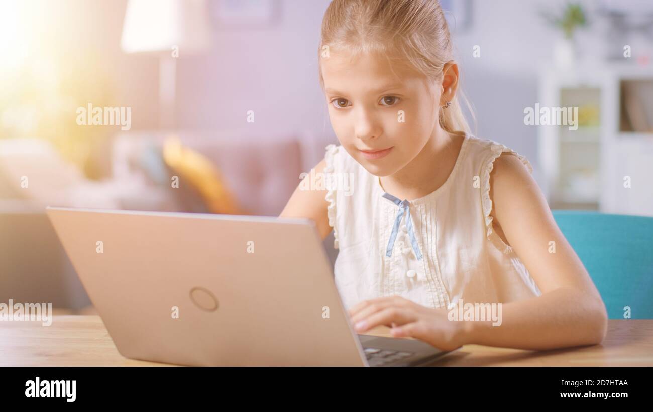 Mignon petite fille utilise un ordinateur portable tout en étant assis au bureau dans le salon. Enfant faisant des devoirs sur ordinateur, navigue par Internet et montres Banque D'Images