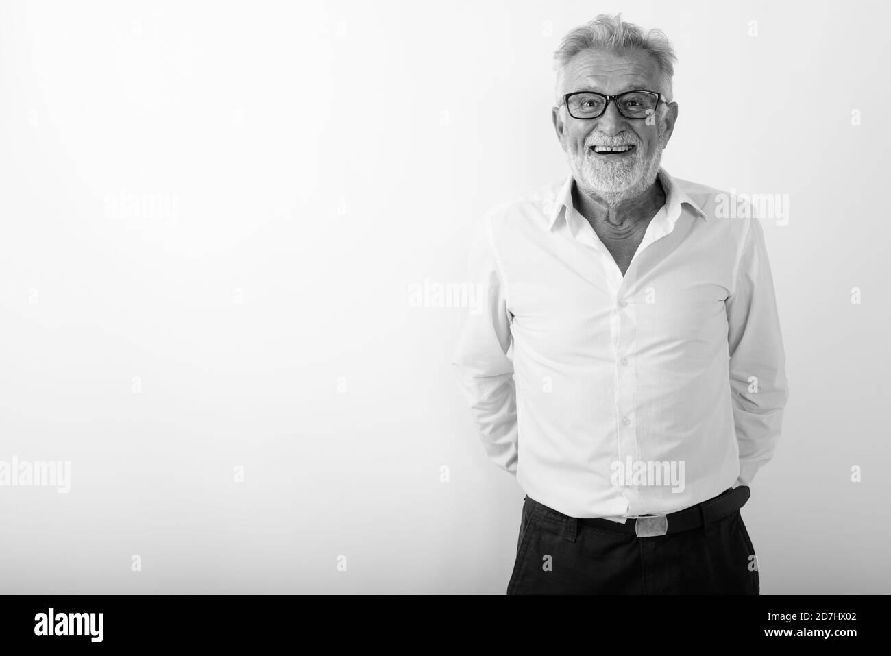 Studio shot of happy senior man smiling barbus avec les mains derrière le dos en portant des lunettes contre fond blanc Banque D'Images