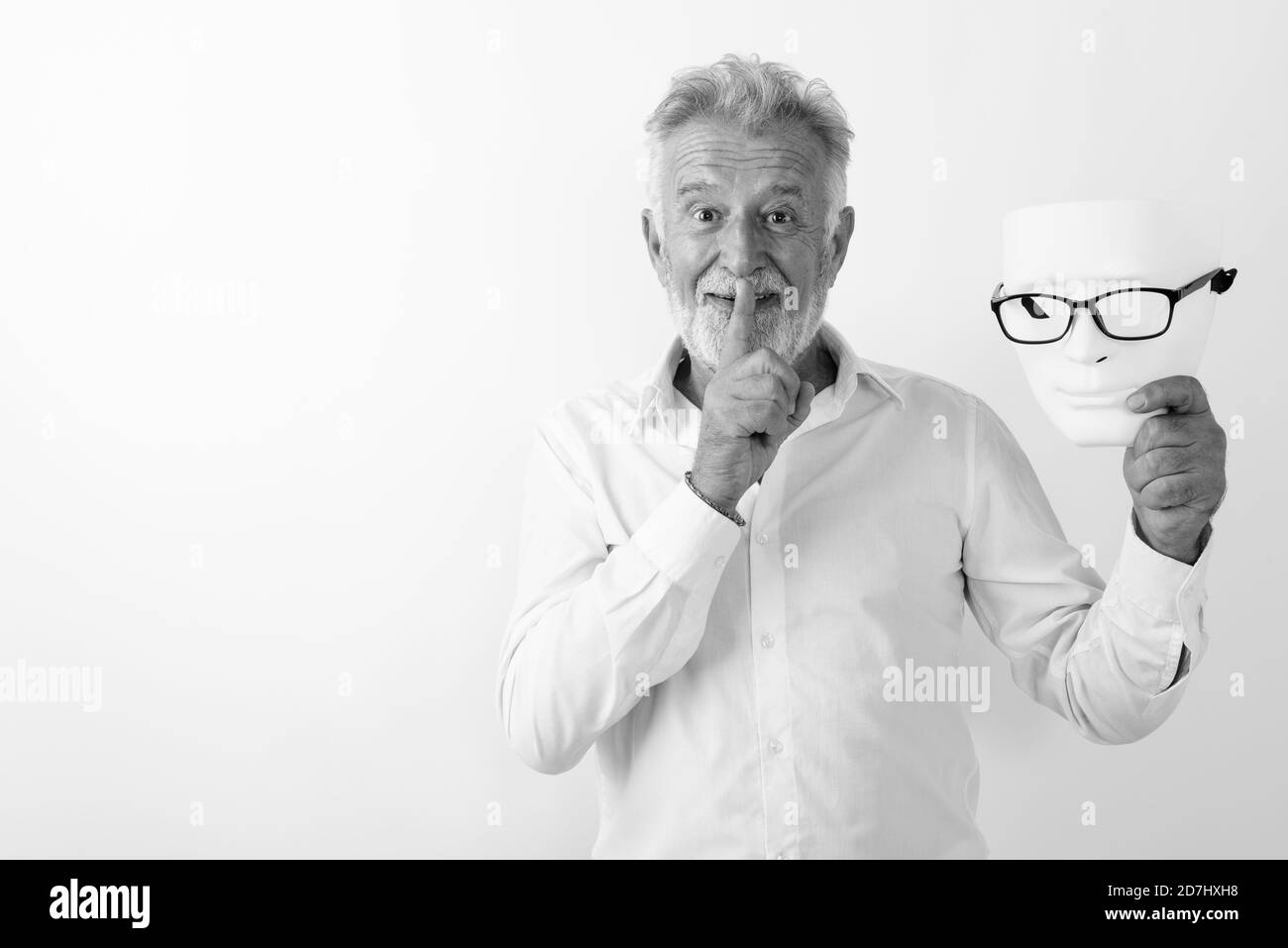 Studio shot of happy senior man smiling barbu avec le doigt sur les lèvres tout en maintenant le masque blanc avec des lunettes contre fond blanc Banque D'Images