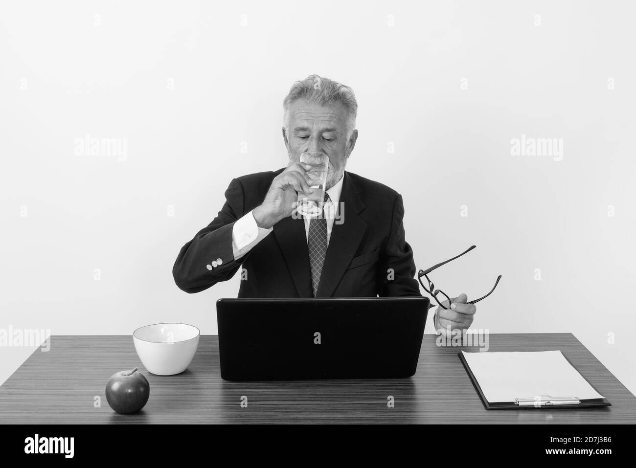 Photo de studio d'un homme d'affaires barbu de haut niveau tenant des lunettes de vue pendant de l'eau potable et de l'ordinateur portable Banque D'Images