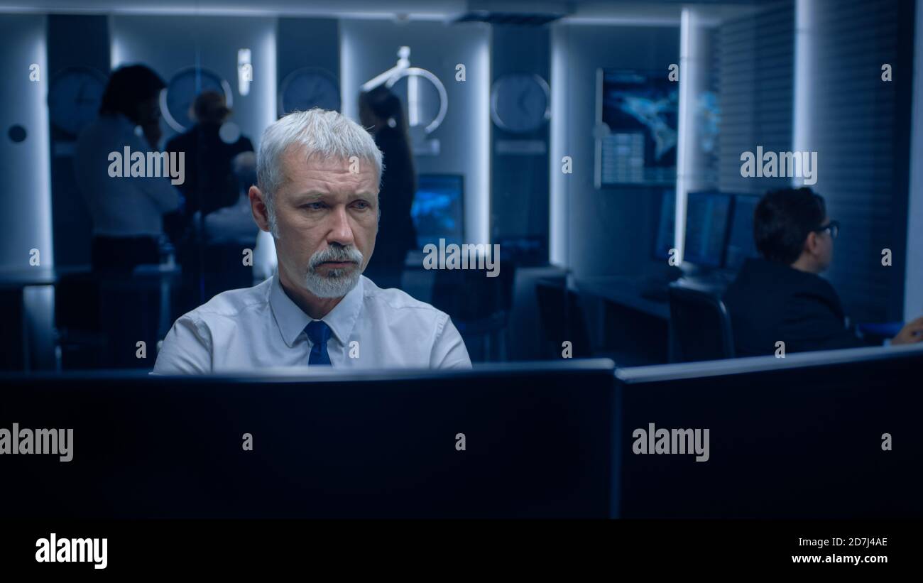 Portrait de l'expert principal concerné travaillant sur l'ordinateur dans la salle de contrôle de la surveillance avec des équipes qui résolvent des problèmes en arrière-plan. Banque D'Images