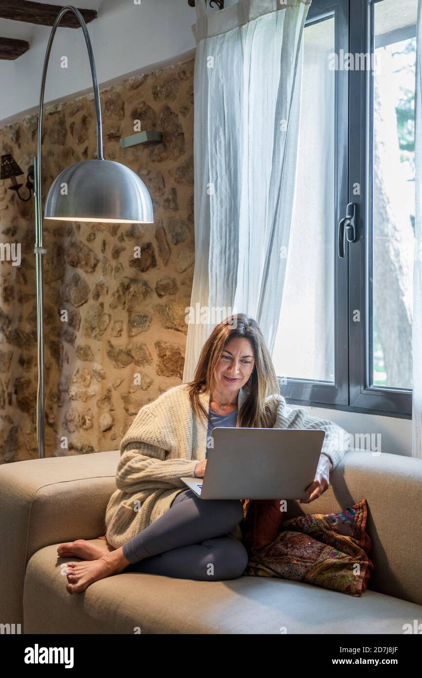 Femme travaillant sur un ordinateur portable tout en étant assise sur un canapé près de la fenêtre à la maison Banque D'Images