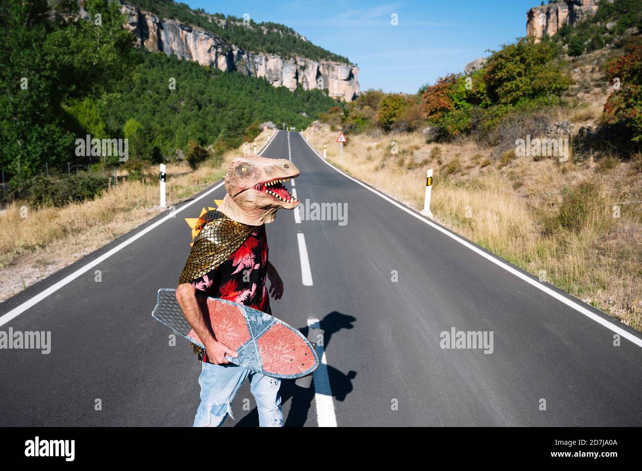 Homme portant un masque de dinosaure tenant une planche à roulettes tout en marchant sur la route Banque D'Images
