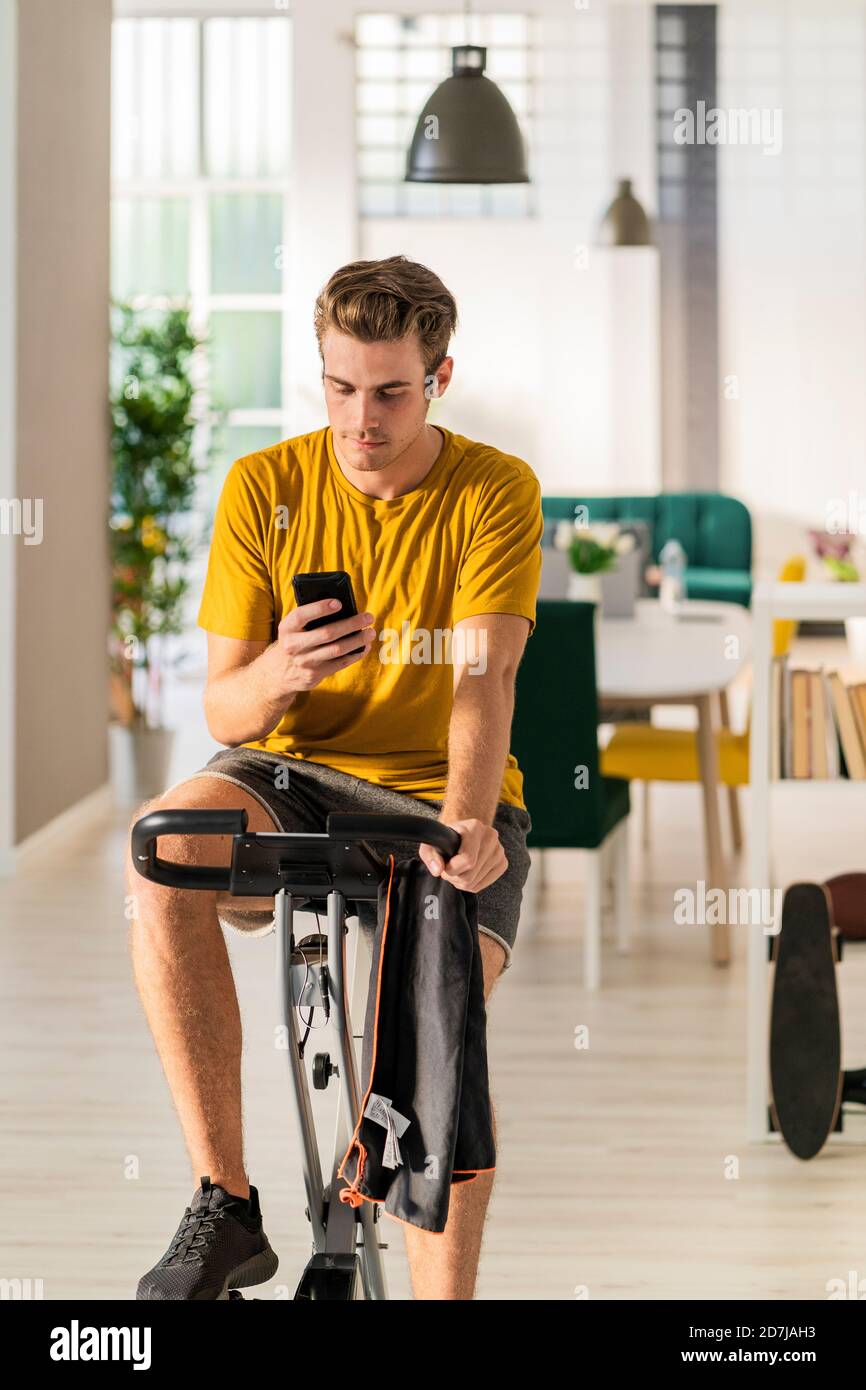 Jeune homme utilisant le téléphone portable tout en faisant du vélo sur l'équipement d'exercice à la maison Banque D'Images