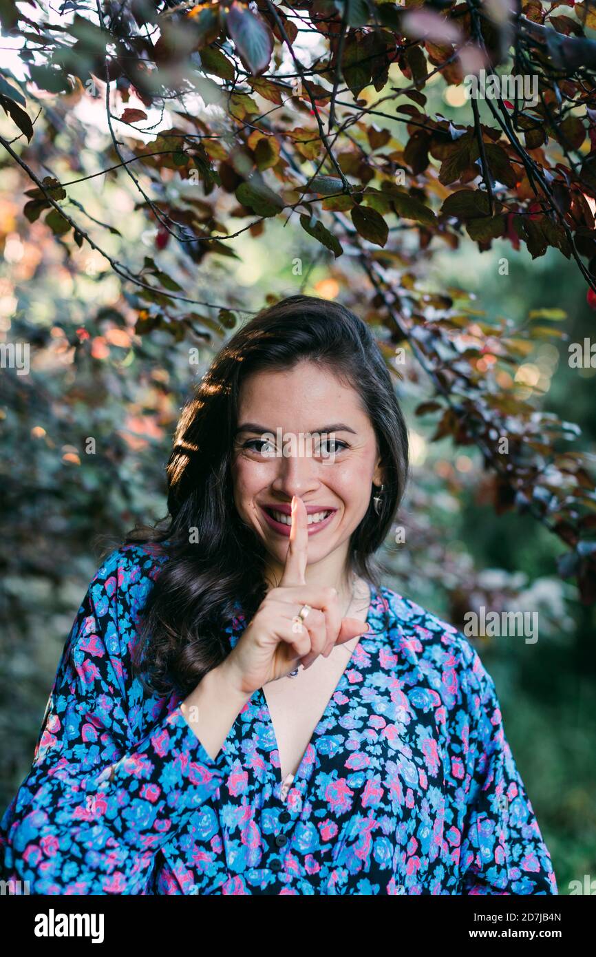 Gros plan d'une jeune femme souriante avec le doigt sur les lèvres debout contre les plantes dans le parc Banque D'Images