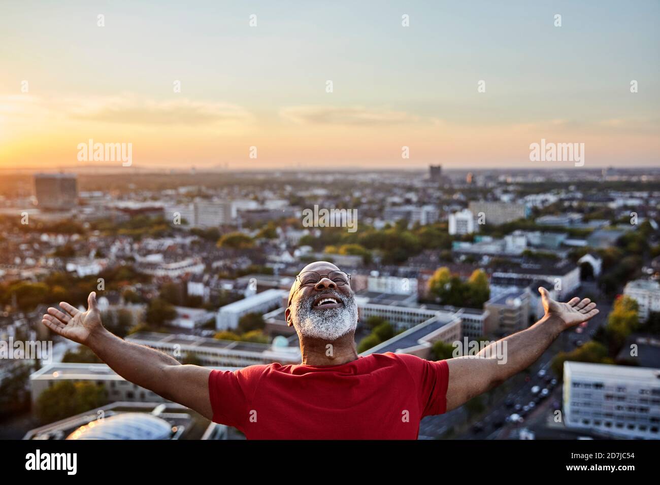 Homme à barbe heureux avec les bras étirés tout en se tenant debout sur le bâtiment terrasse en ville au coucher du soleil Banque D'Images