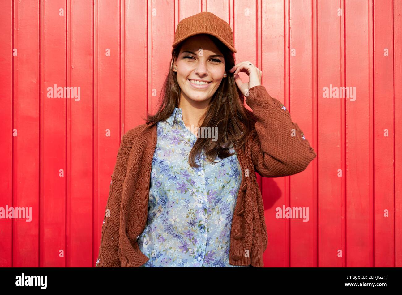 Femme souriante avec un chapeau debout contre une porte en métal rouge Banque D'Images