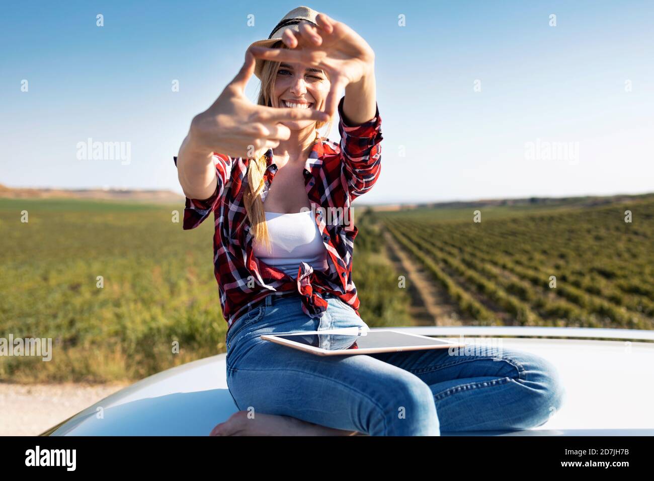 Jeune femme souriante montrant le cadre du doigt tout en étant assise sur la voiture toit Banque D'Images