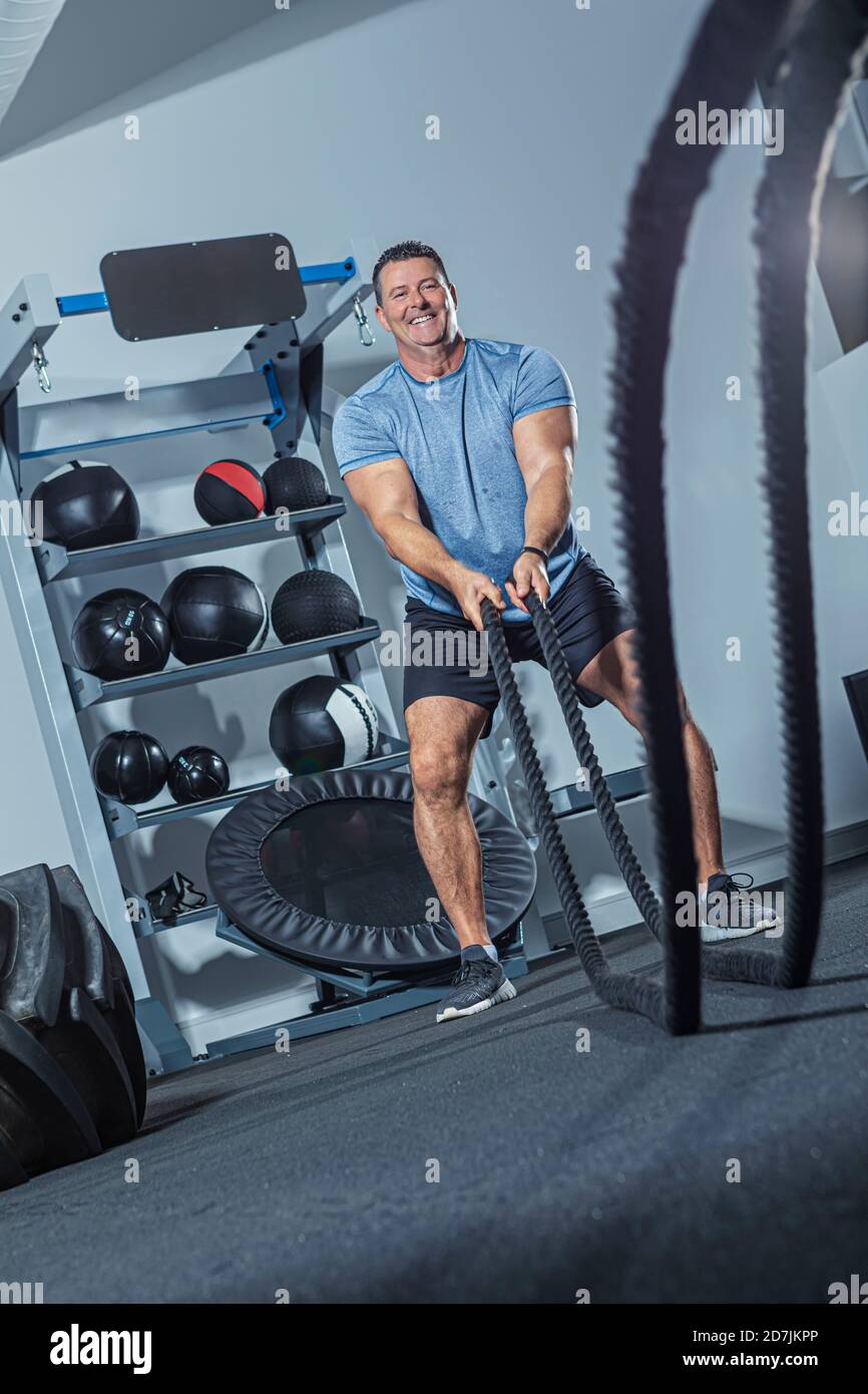 Homme mûr souriant s'exerçant avec corde de bataille dans la salle de gym Banque D'Images