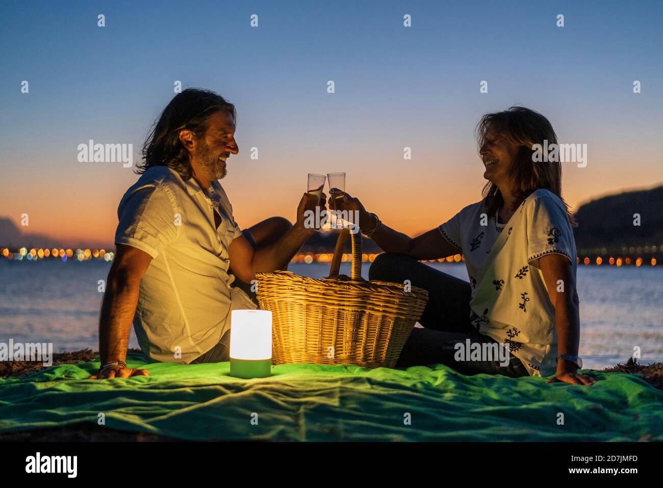 Couple toaster les boissons tout en étant assis près du panier à la plage contre ciel dégagé au coucher du soleil Banque D'Images