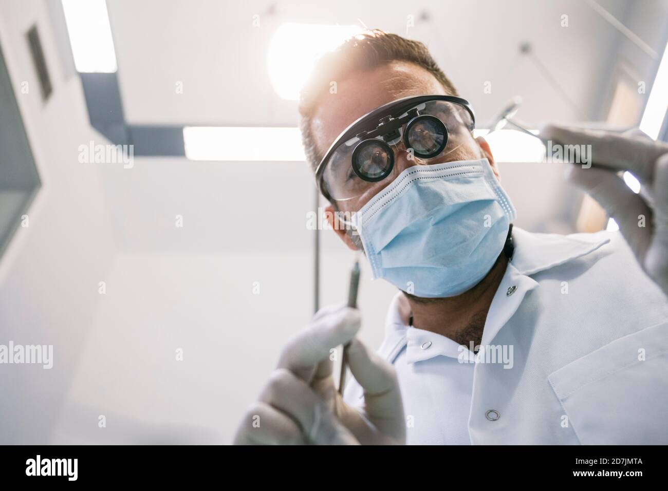 Dentiste de sexe masculin dans des loupes chirurgicales et un masque avec instruments médicaux pendant le traitement en clinique Banque D'Images