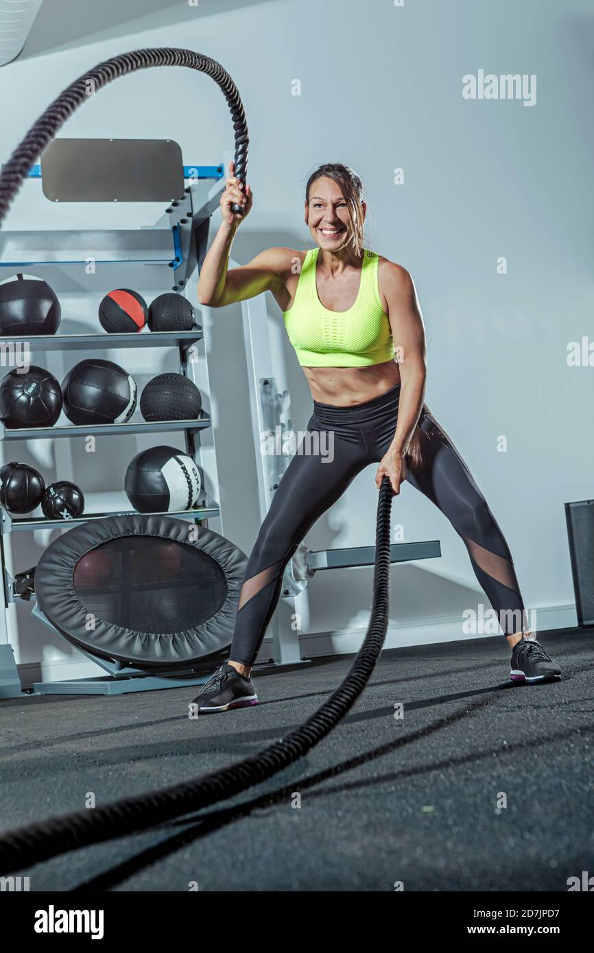 Femme mûre souriante s'exerçant avec corde de bataille dans la salle de gym Banque D'Images