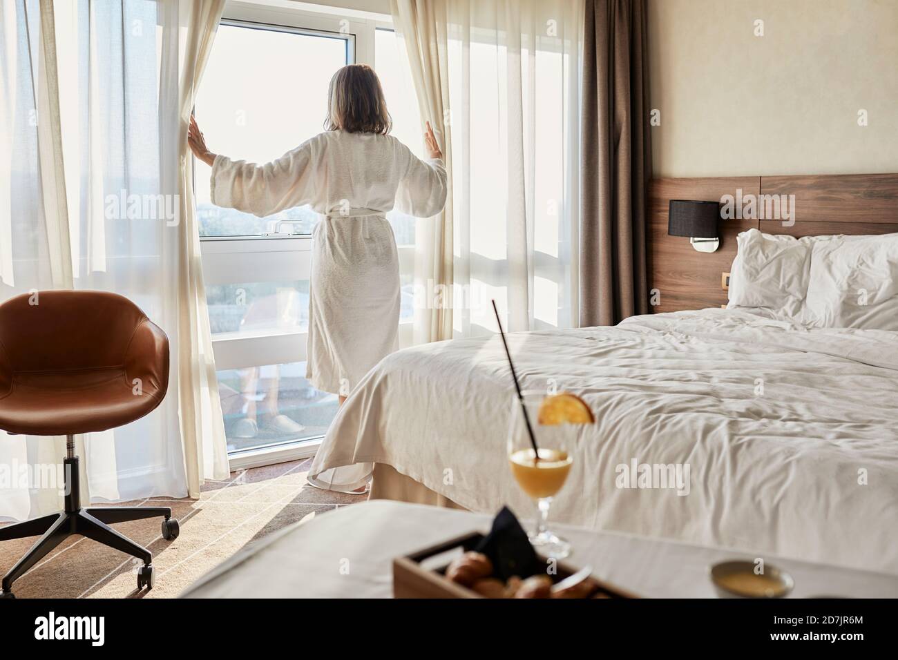Femme âgée à la retraite ouvrant le rideau tout en regardant par la fenêtre à chambre d'hôtel de luxe Banque D'Images