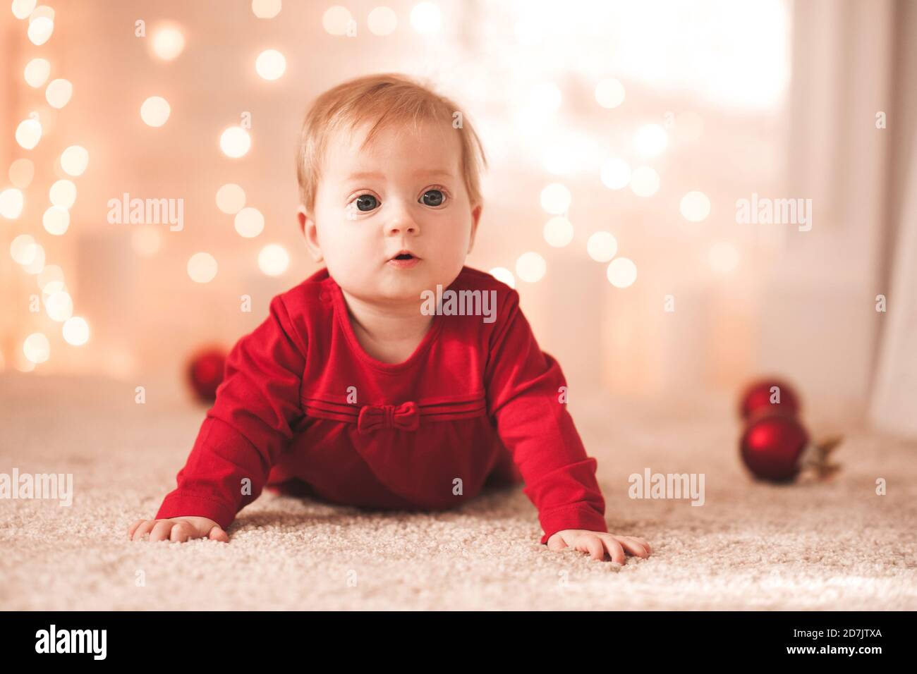 Mignon bébé fille de moins de 1 an portant robe rouge rampant sur le sol avec décor de Noël sur des lumières brillantes gros plan. Saison des vacances d'hiver. Childhoo Banque D'Images