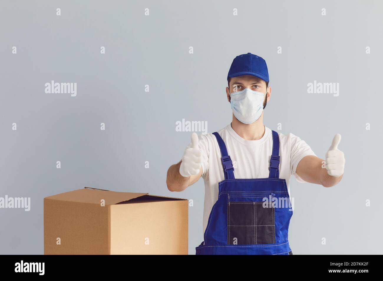 Un messager masculin en uniforme, un masque médical et des gants lève les pouces sur fond de mur gris. Banque D'Images