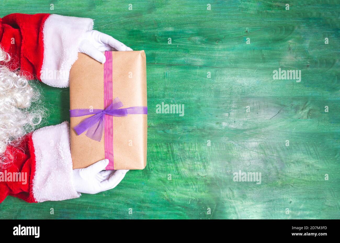 Détail des bras et des mains avec les gants du Père Noël et barbe offrant un cadeau avec des rubans sur un bois vert Arrière-plan à Noël Banque D'Images
