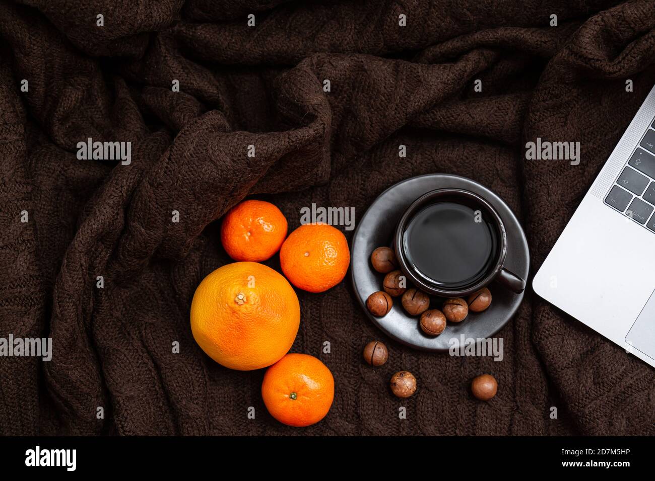Ambiance chaleureuse. Ordinateur portable, tasse de café avec mandarines et noix sur fond de couverture tricoté marron Banque D'Images
