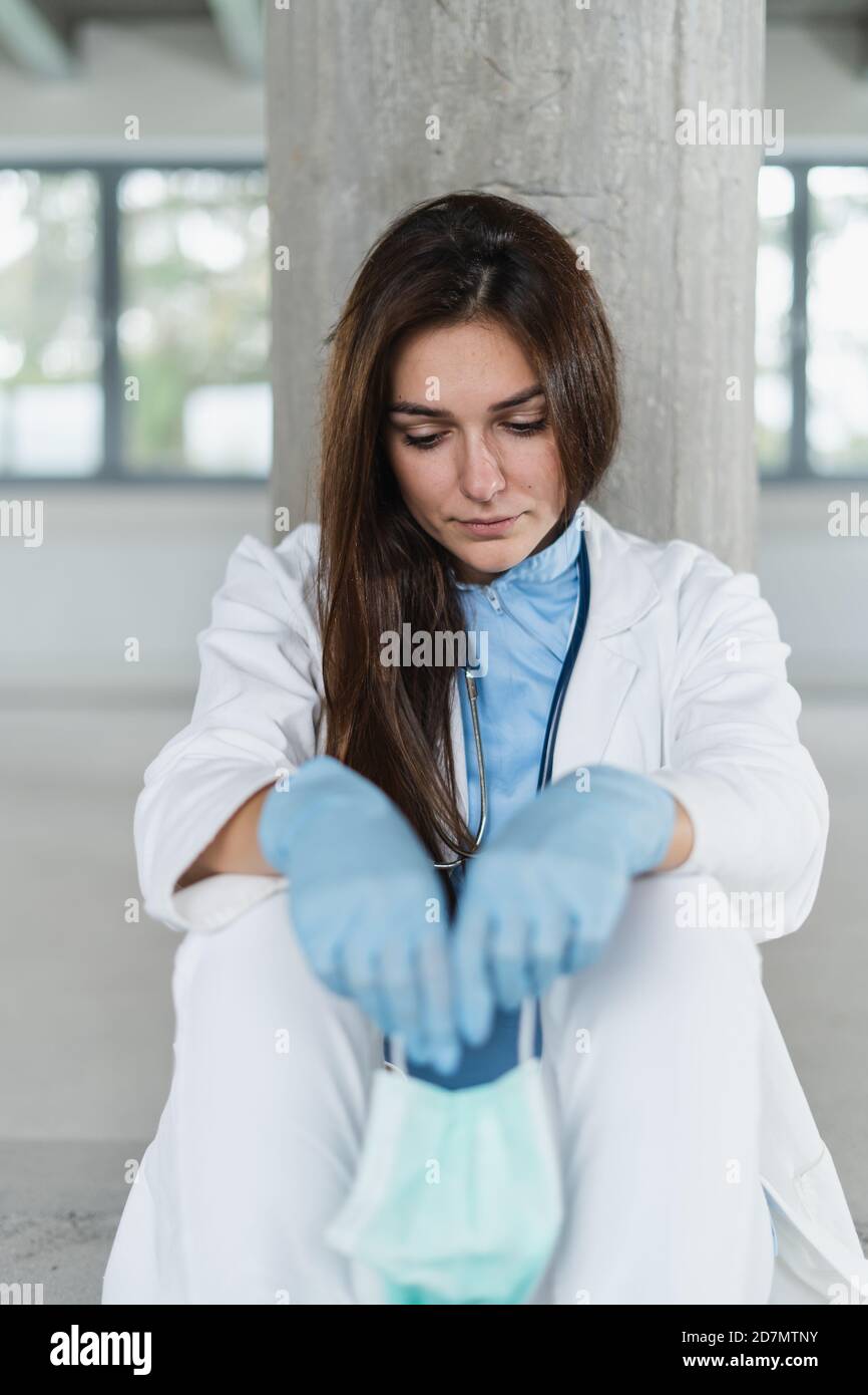 Jeune femme triste et désespéré médecin tenant un masque protecteur assis sur le sol à l'hôpital après le quart de travail. Coronavirus covid-19 concept. Banque D'Images