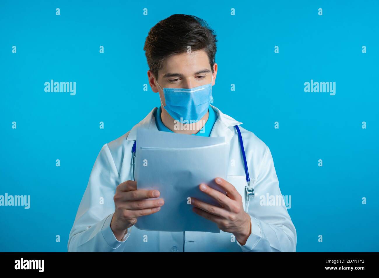 Homme satisfait dans un pelage médical professionnel et masque tenant des dossiers papiers isolés sur fond bleu. Il nod sa tête approvamment au diagnostic docteur Banque D'Images
