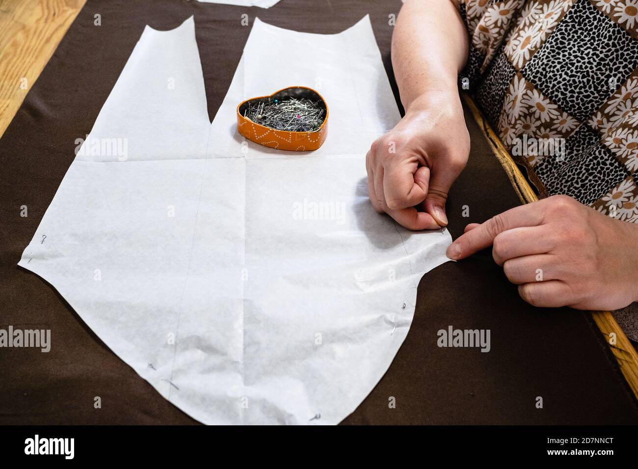 l'application sur mesure fixe la disposition en papier de la robe au tissu brun par des épingles sur une table en bois à la maison Banque D'Images