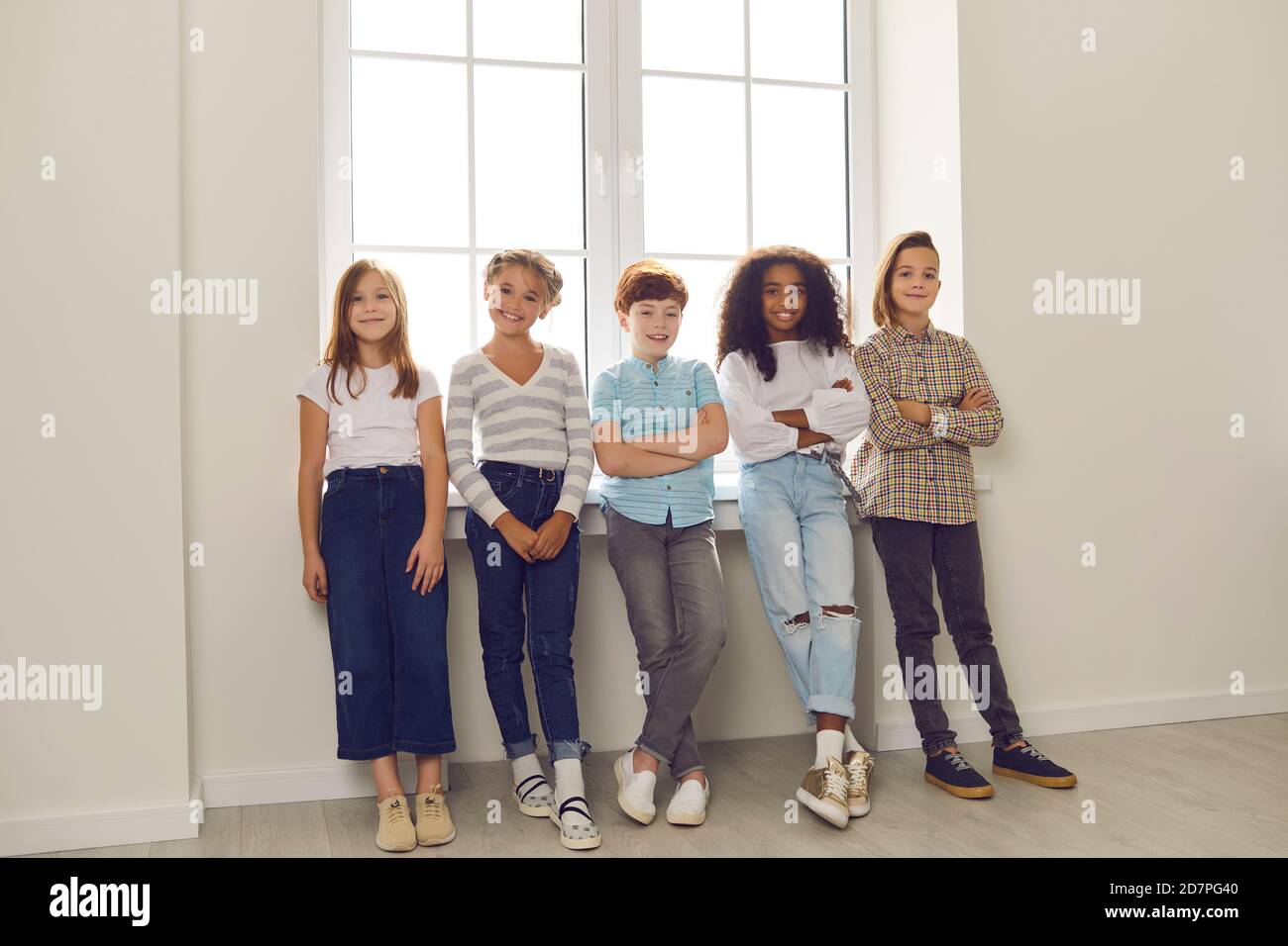 Groupe d'enfants heureux et divers debout près de la fenêtre, regardant l'appareil photo et souriant Banque D'Images
