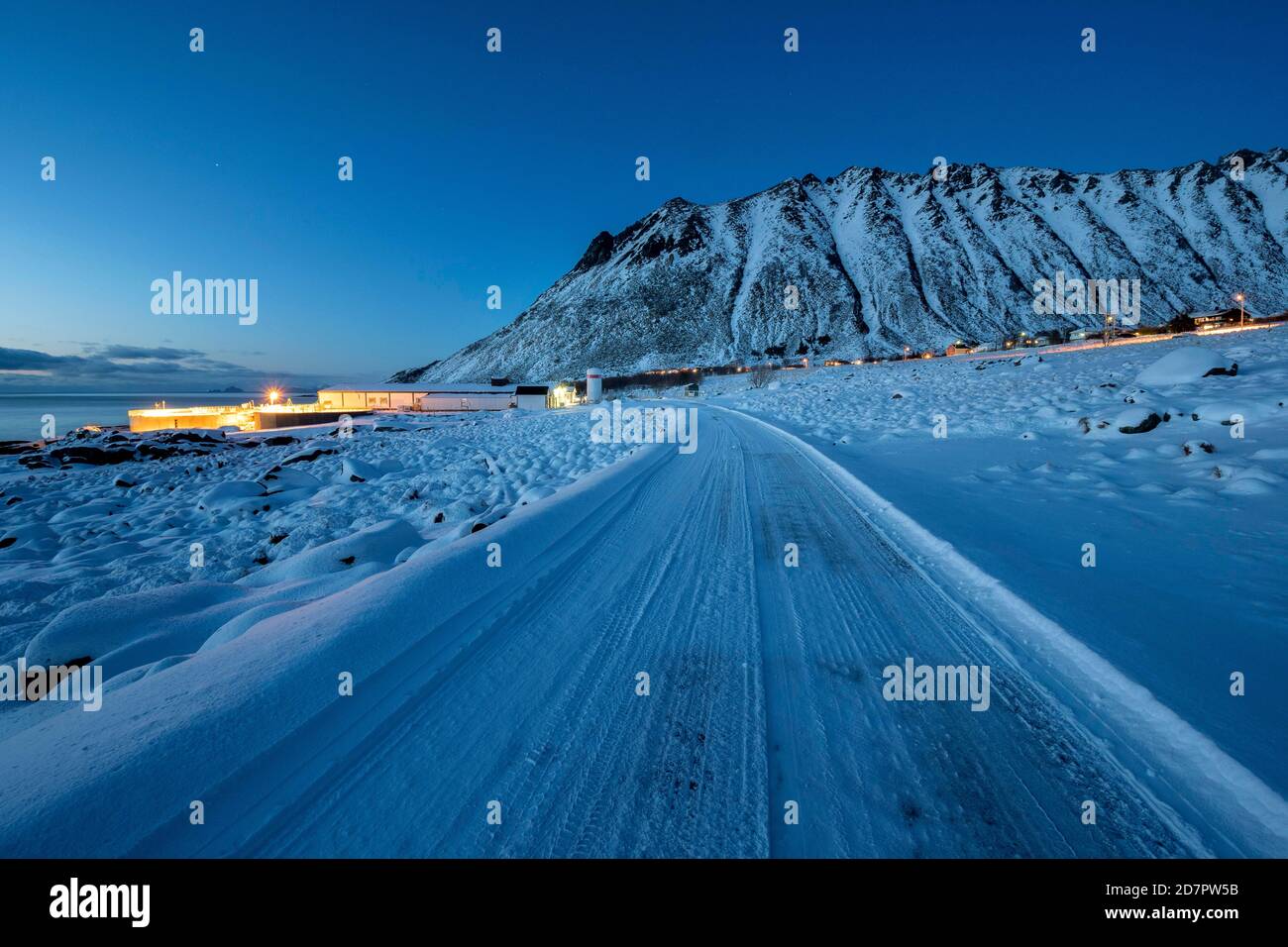 Route enneigée glacée, village éclairé au crépuscule, bâtiments résidentiels et industriels, Nordland, Lofoten, Norvège Banque D'Images