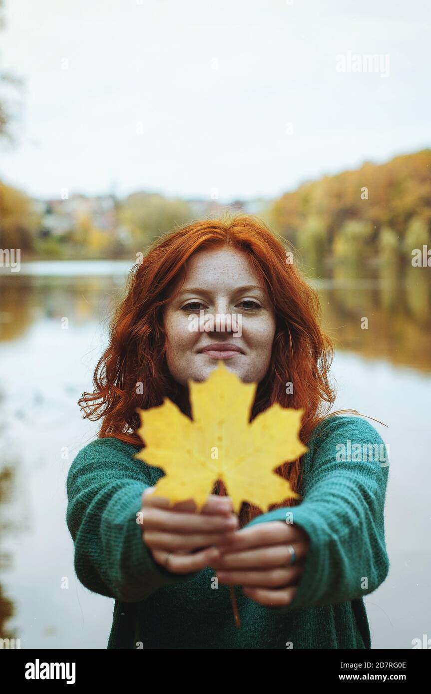 Portrait amusant d'une jeune femme à tête rouge avec une feuille d'automne jaune dans ses mains dans le parc. Banque D'Images