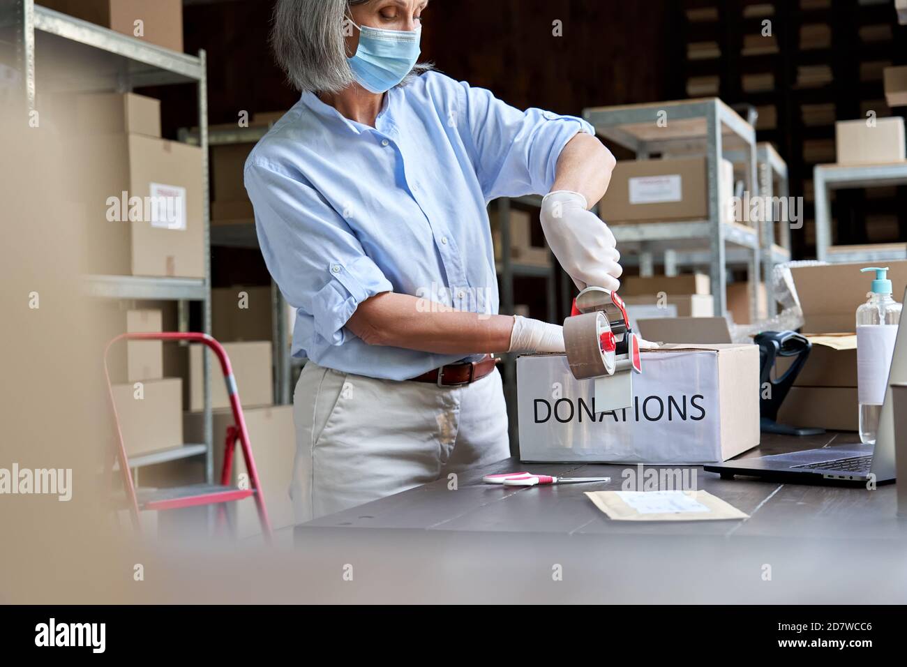 Une femme mature travaillant dans un entrepôt portant un masque d'emballage contenant une boîte de dons. Banque D'Images