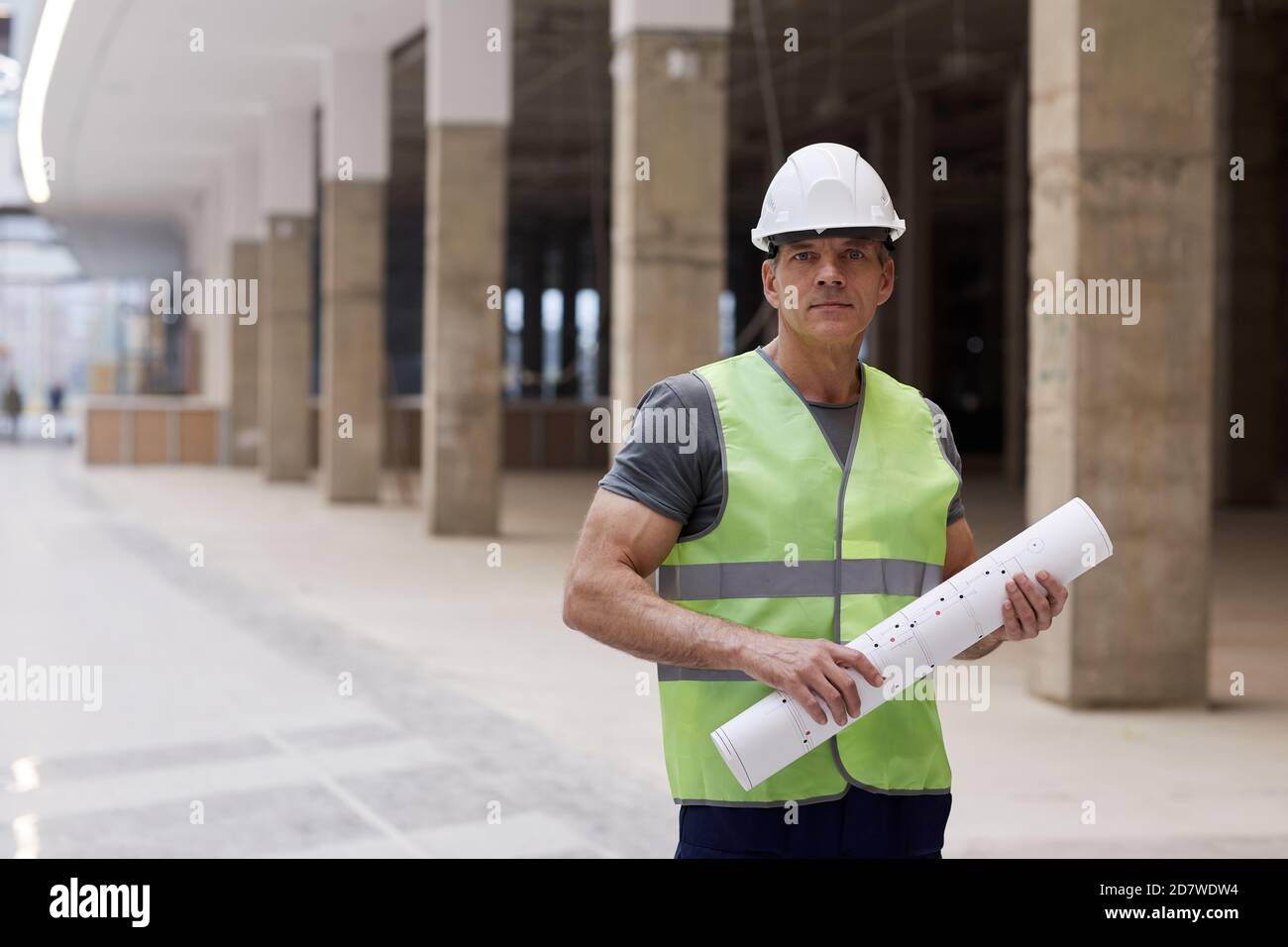 Portrait à la taille de l'ouvrier de construction professionnel tenant des plans et regardant l'appareil photo tout en se tenant dans un immeuble de bureau, espace de copie Banque D'Images