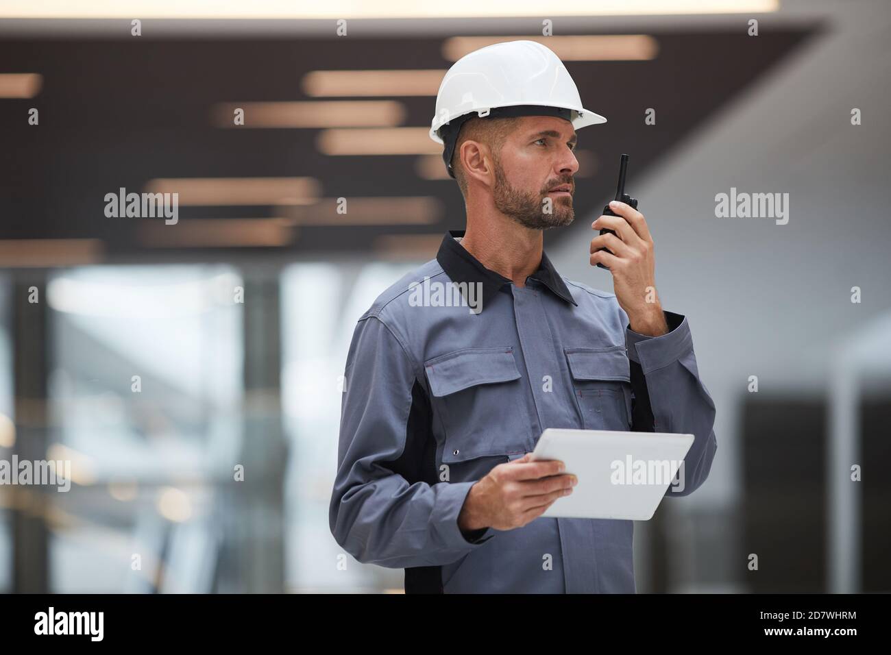 Portrait à la taille d'un beau travailleur mûr parlant par walkie-talkie tout en supervisant le travail sur le chantier ou dans un atelier industriel, espace de copie Banque D'Images
