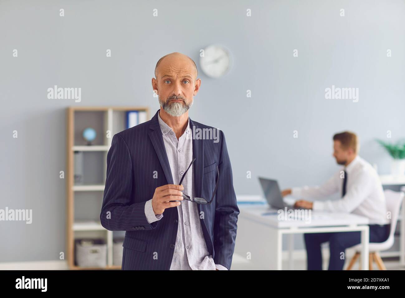 Cadre d'entreprise mature debout au bureau regardant la caméra avec employé travaillant en arrière-plan Banque D'Images