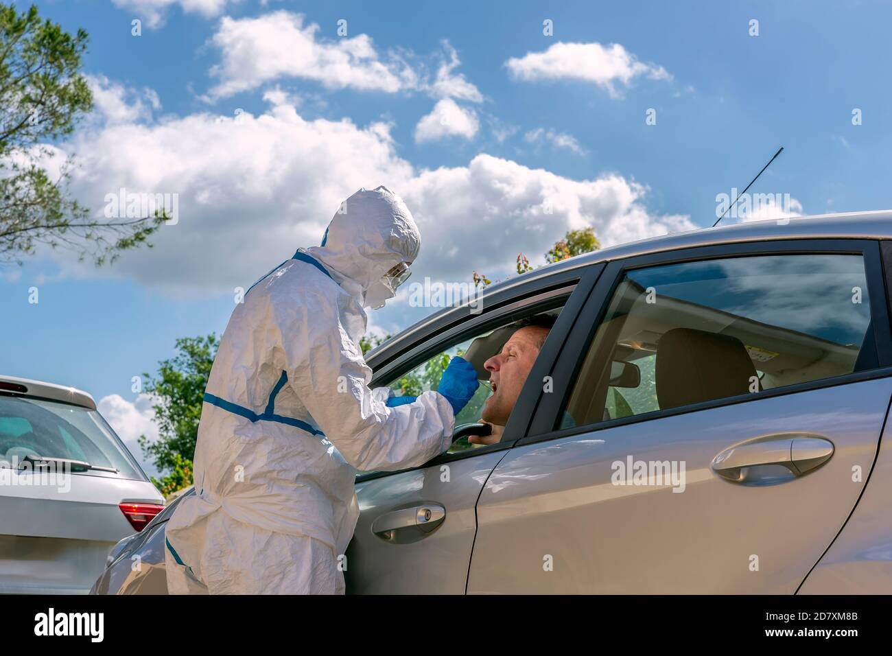 Un professionnel de la santé entièrement couvert d'un costume de protection blanc et de lunettes fait fonctionner un écouvillon pour Covid-19 à un homme assis à l'intérieur de sa voiture Banque D'Images