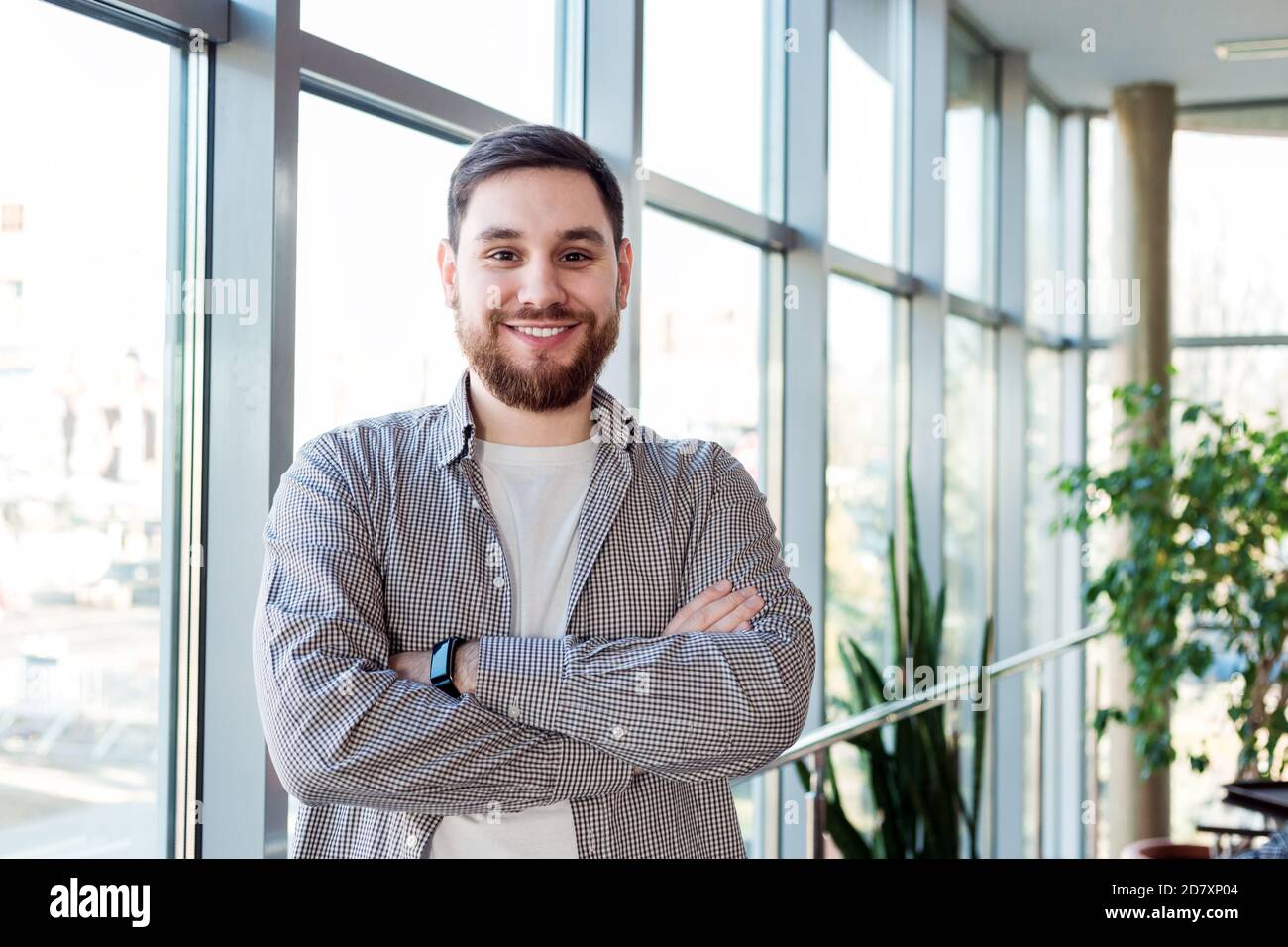 Homme caucasien souriant debout près de la fenêtre pleine longueur dans le bureau seul. Un jeune homme barbu heureux et réussi a croisé les bras. Un homme d'affaires élégant Banque D'Images