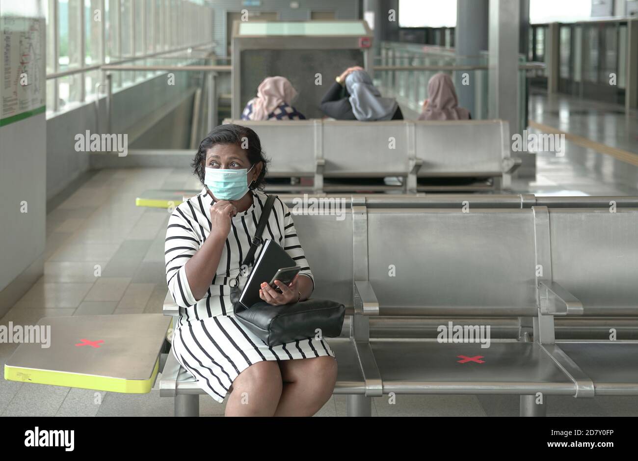 Femme portant un masque de visage assis à la station de métro attendant entraînez-vous avec d'autres personnes en arrière-plan Banque D'Images