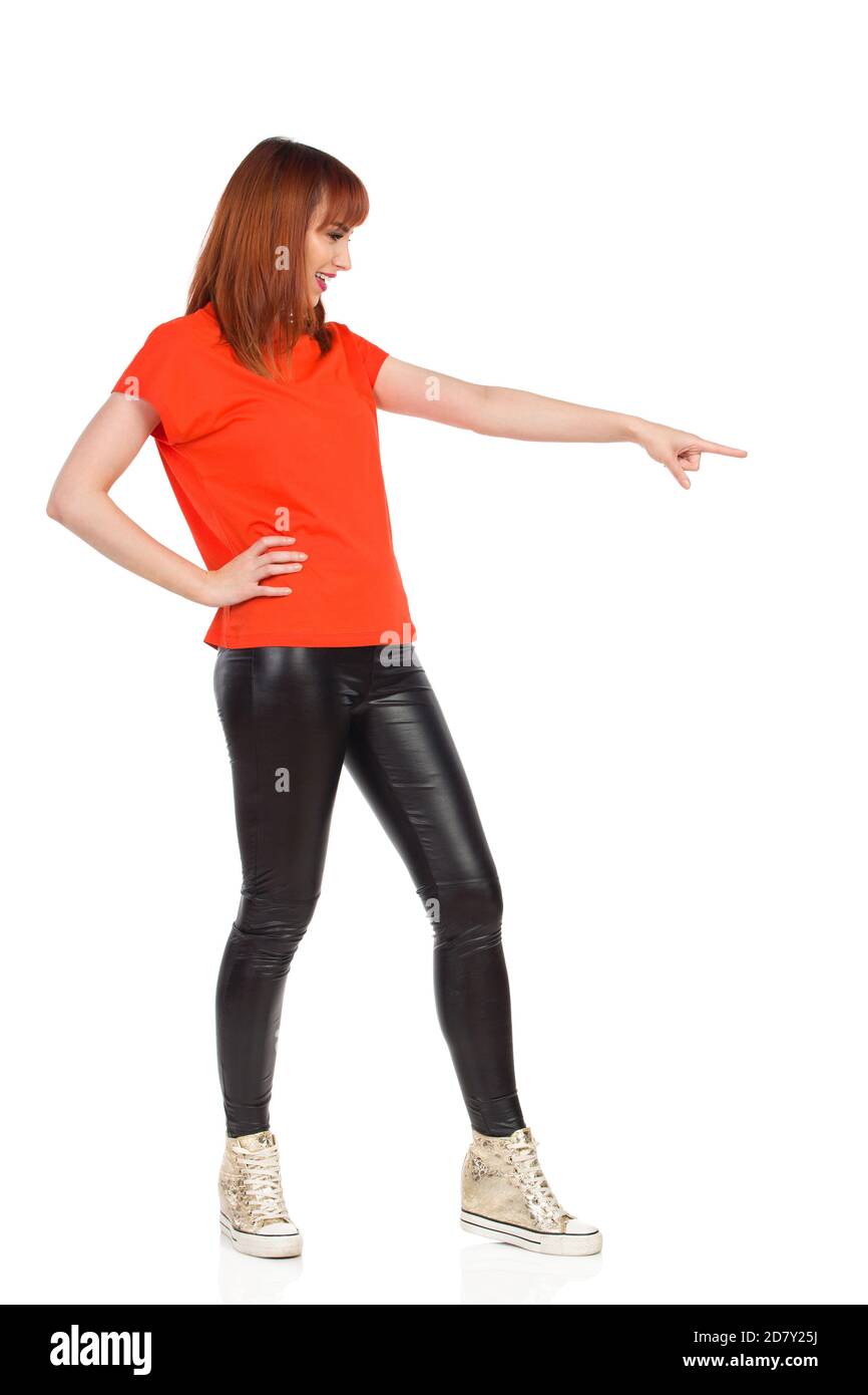 Décontracté jeune femme en t-shirt orange, pantalon noir et baskets or brillant est debout regardant loin et pointant sur le côté. Plein studio tourné i Banque D'Images