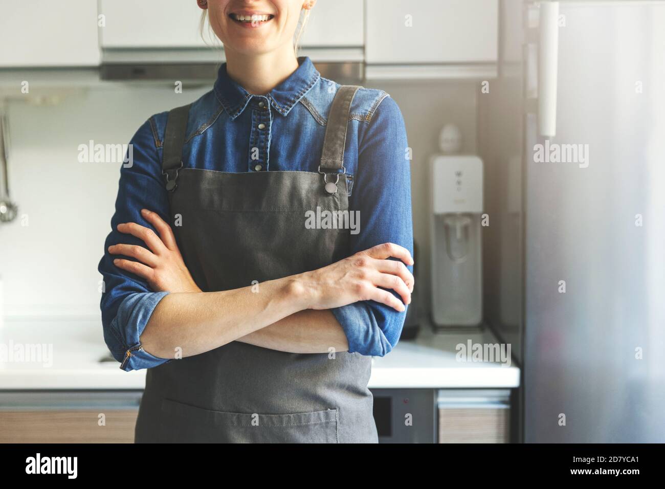 jeune femme souriante avec tablier debout dans la cuisine maison avec bras croisés Banque D'Images