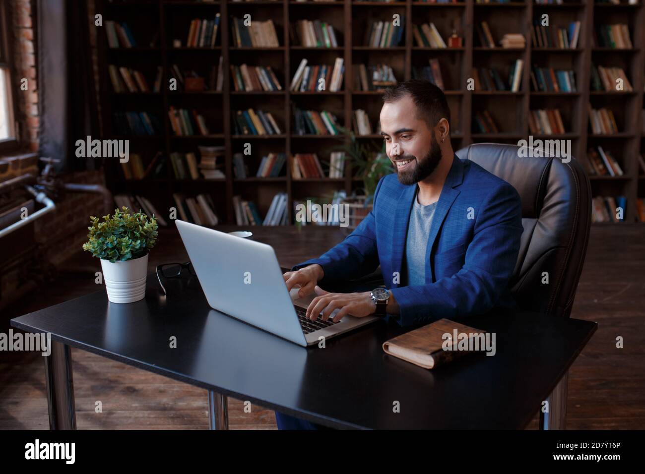Un jeune homme d'affaires travaille sur un ordinateur portable dans un bureau luxueux de la bibliothèque. Un homme barbu, gai dans un costume bleu à un bureau est de taper sur un clavier Banque D'Images