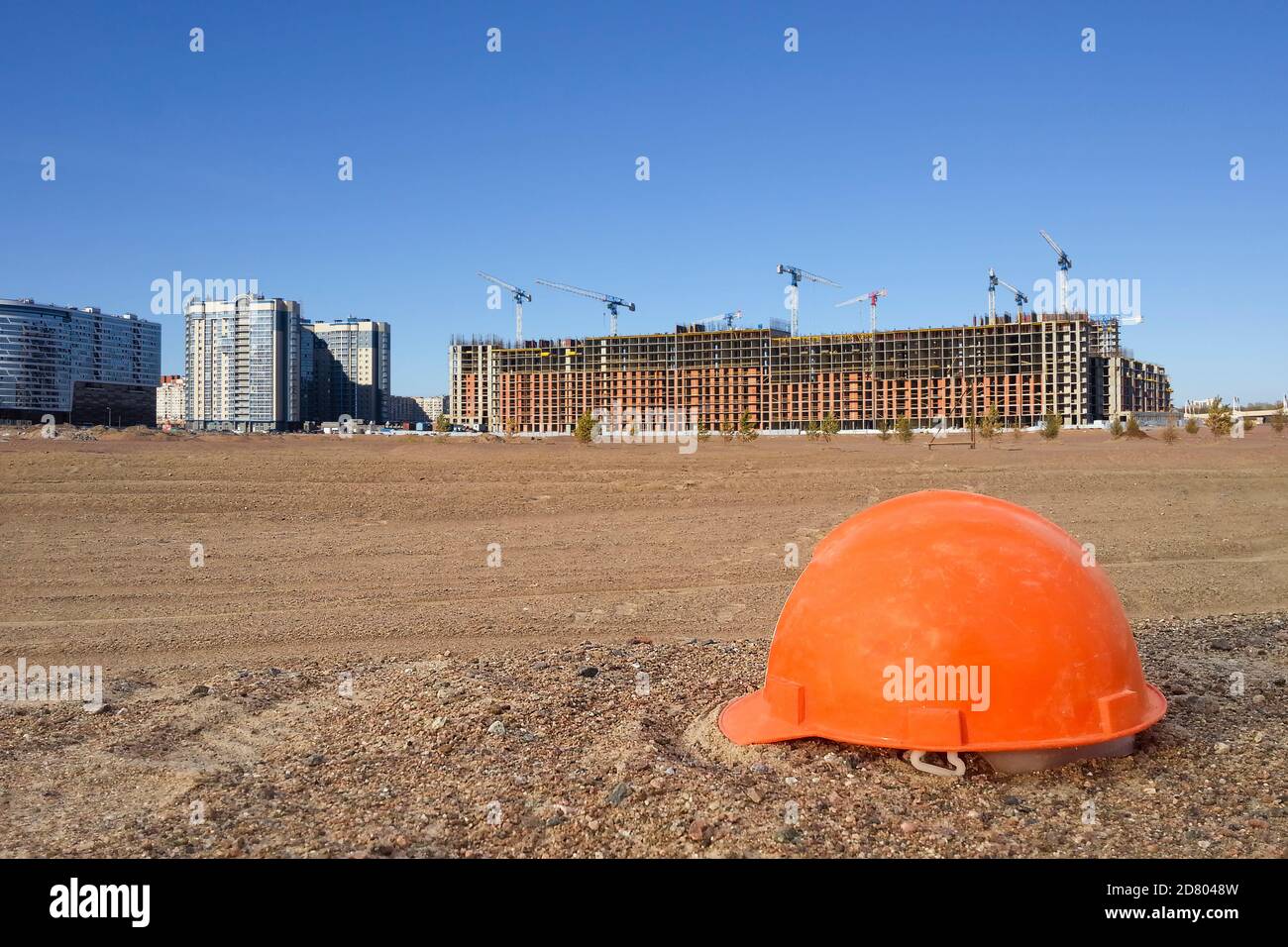 Un casque de sécurité de couleur orange sur un sol sablonneux sur le fond d'un chantier de construction, de bâtiments et de grues Banque D'Images