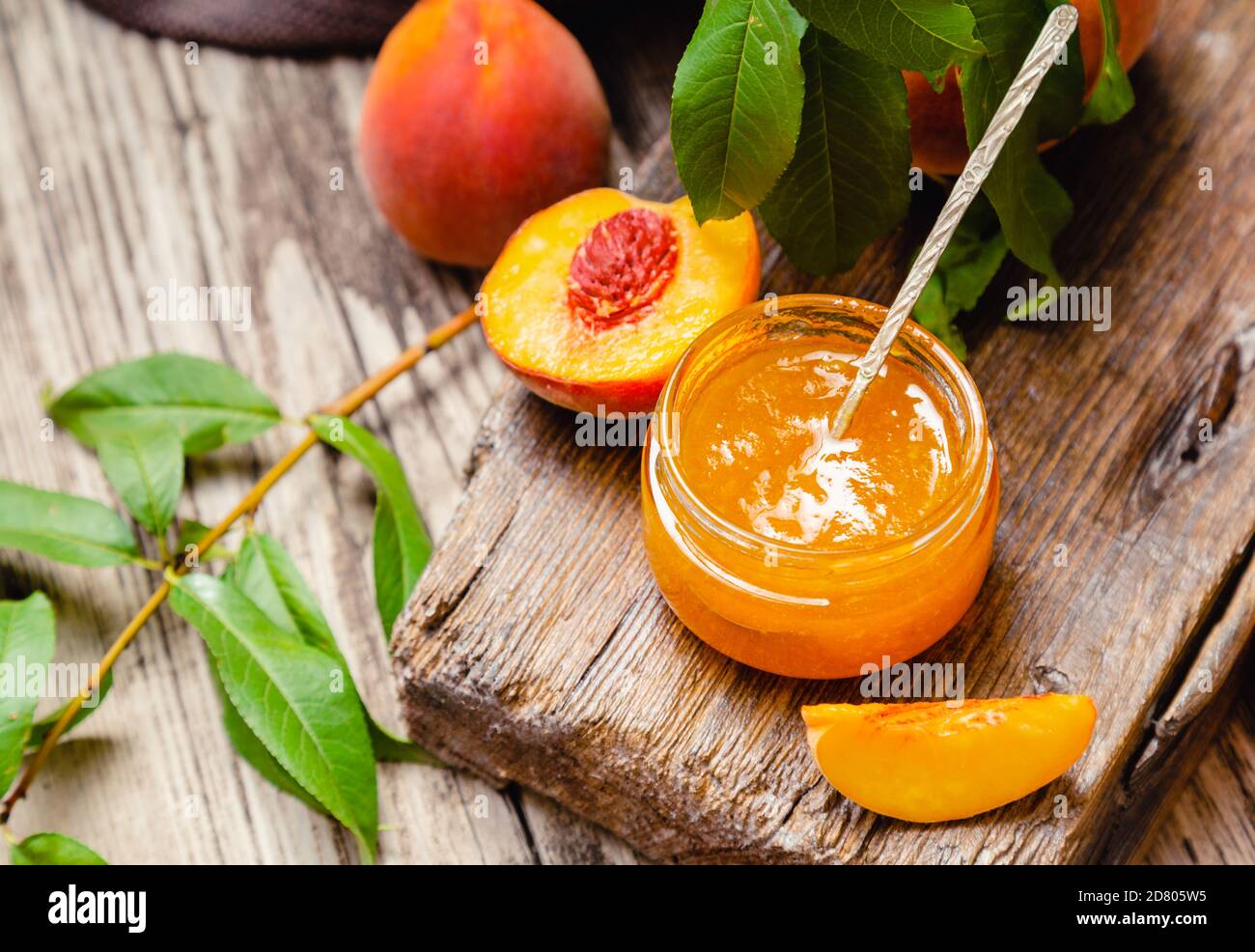 Confiture de pêche dans un pot en verre avec des quartiers et des fruits de pêche entiers, des feuilles d'arbre de pêche. Confiture de pêche sur une table en bois avec une cuillère. Banque D'Images