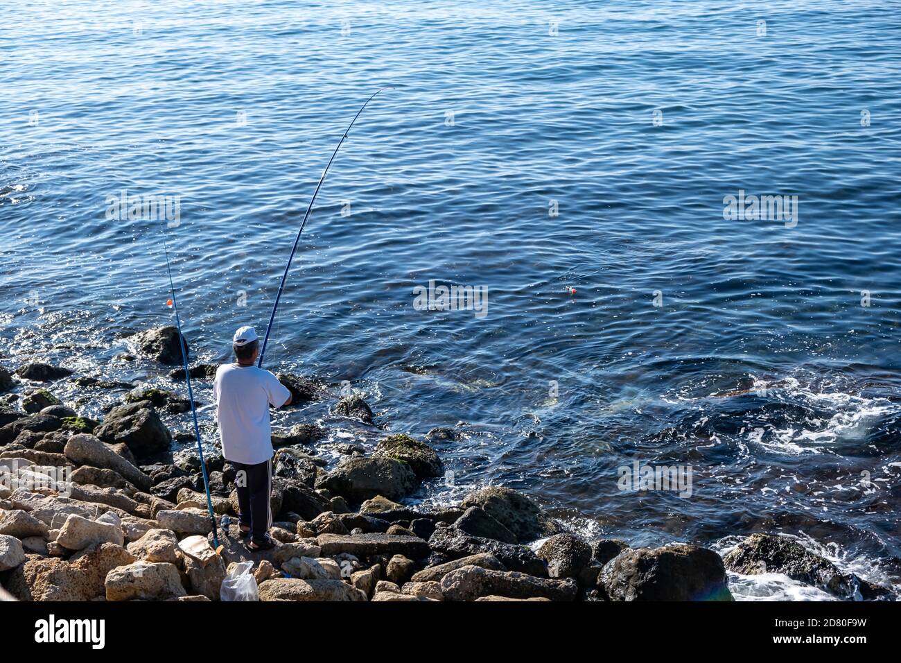 Pêcheur debout sur des rochers avec deux cannes à pêche un jour ensoleillé. Chapeau blanc pour la protection du soleil, les loisirs et la nourriture gratuite. Fond bleu de mer agité, au-dessus Banque D'Images