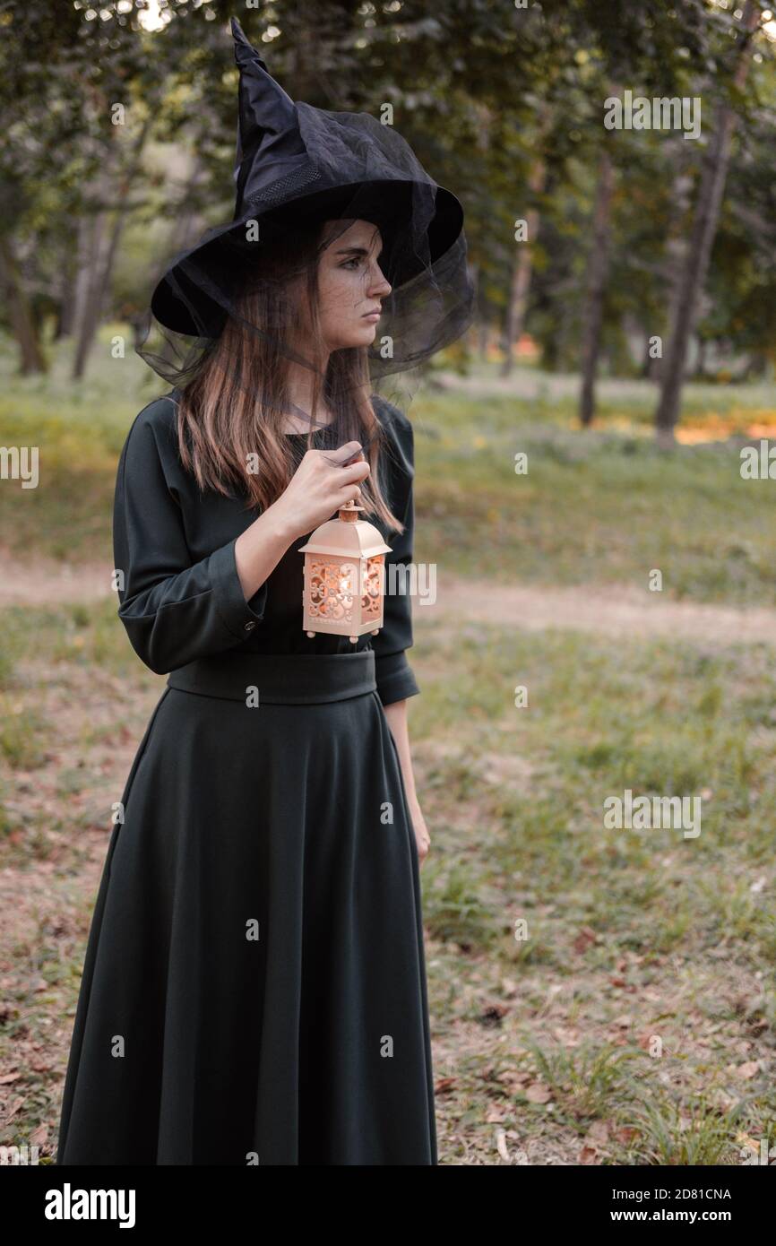 Une jeune femme en robe sombre et chapeau de sorcière tient une lanterne avec des bougies dans ses mains et illumine la forêt. Costume de fête d'Halloween. Garez-vous avec l'autom Banque D'Images