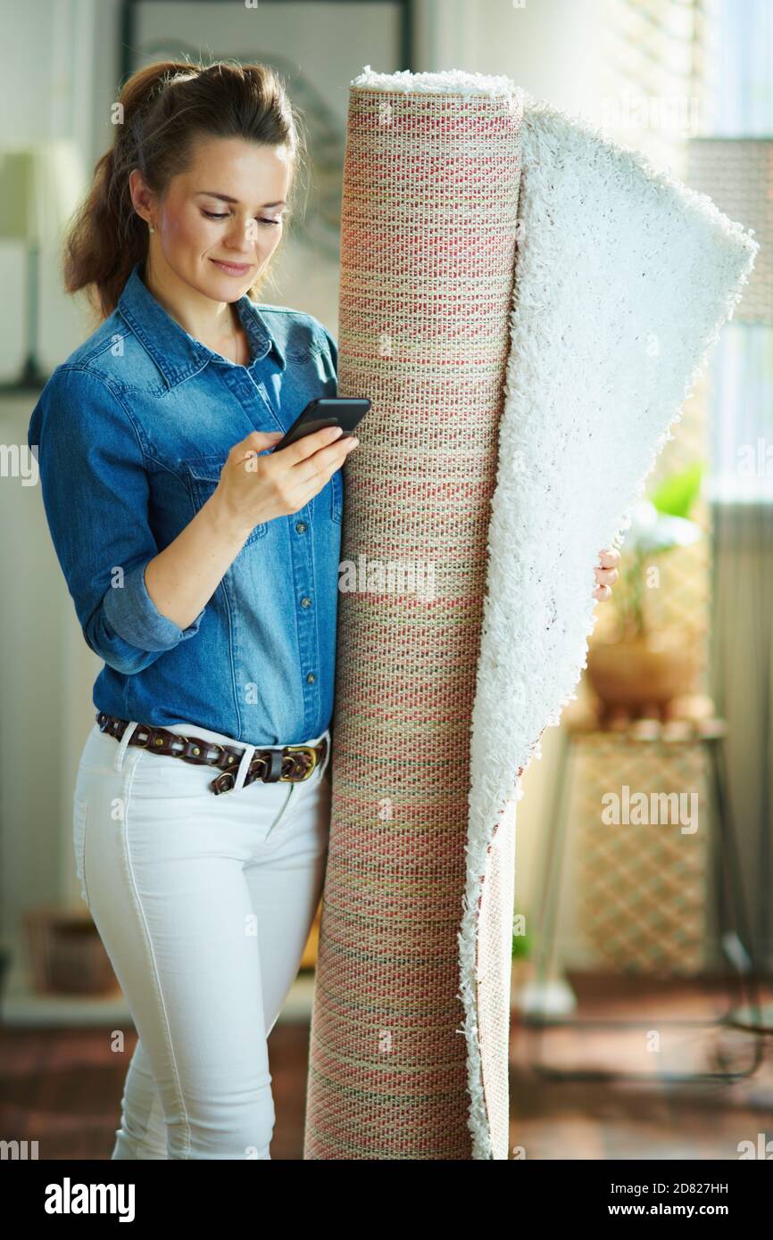 femme au foyer moderne de 40 ans, chemise jean et blanc pantalon avec tapis  blanc à la recherche du service de nettoyage de tapis un smartphone à la  maison moderne dans Photo