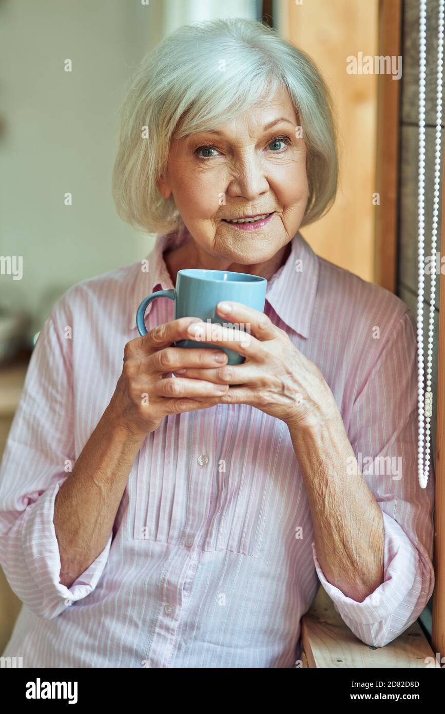 Gros plan de la belle femme aînée souriante se reposant à la maison tout en tenant une boisson chaude dans la cuisine. Concept de style de vie domestique Banque D'Images