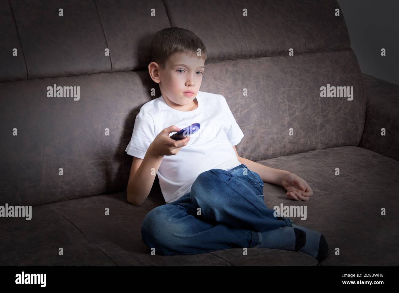 Un joli garçon est assis sur le canapé à l'aide de la télécommande, l'enfant regarde la télévision. Le concept des loisirs pour les enfants, l'impact de la télévision Banque D'Images