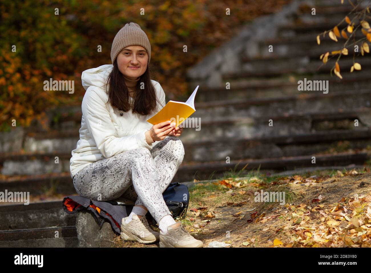 Bonne jolie brune femme assise dans le parc lisant le livre lors d'une belle journée ensoleillée d'automne. Humeur méditative. Banque D'Images