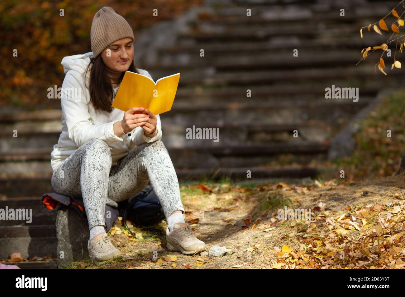 Jolie femme brunette assise dans le parc lisant le livre lors d'une belle journée ensoleillée d'automne. Femme portant un bonnet tricoté, veste chaude. Humeur méditative Banque D'Images