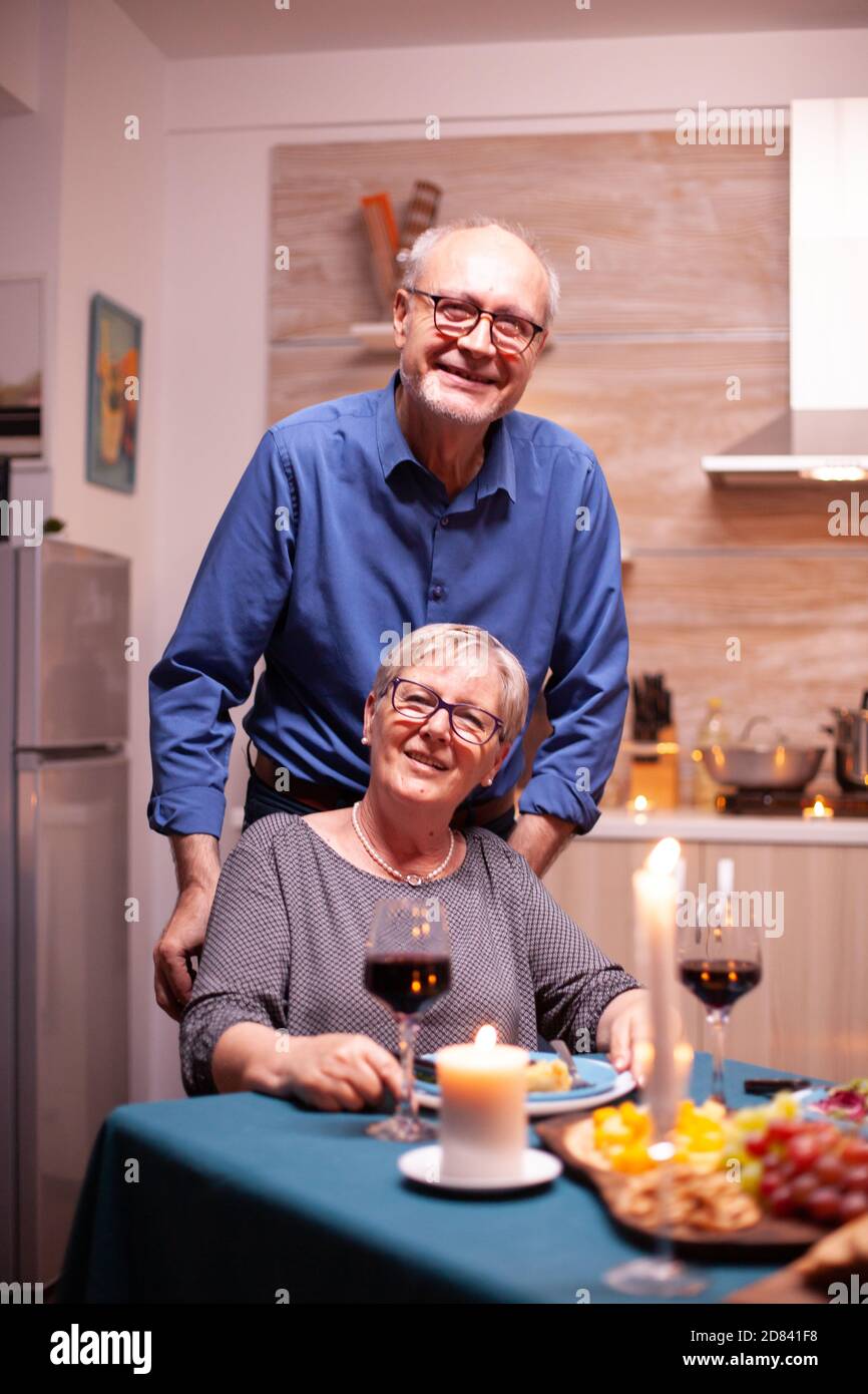 Femme handicapée en fauteuil roulant et mari regardant la caméra. Joyeux couple senior joyeux dînant ensemble dans la cuisine confortable, en appréciant le repas, célébrant leur anniversaire. Banque D'Images