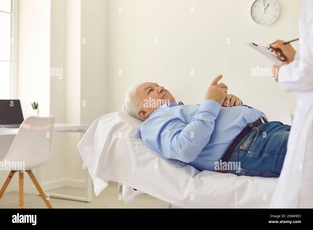 Homme âgé allongé sur le lit dans le bureau de l'hôpital et parlant à médecin qui remplit le dossier de santé Banque D'Images
