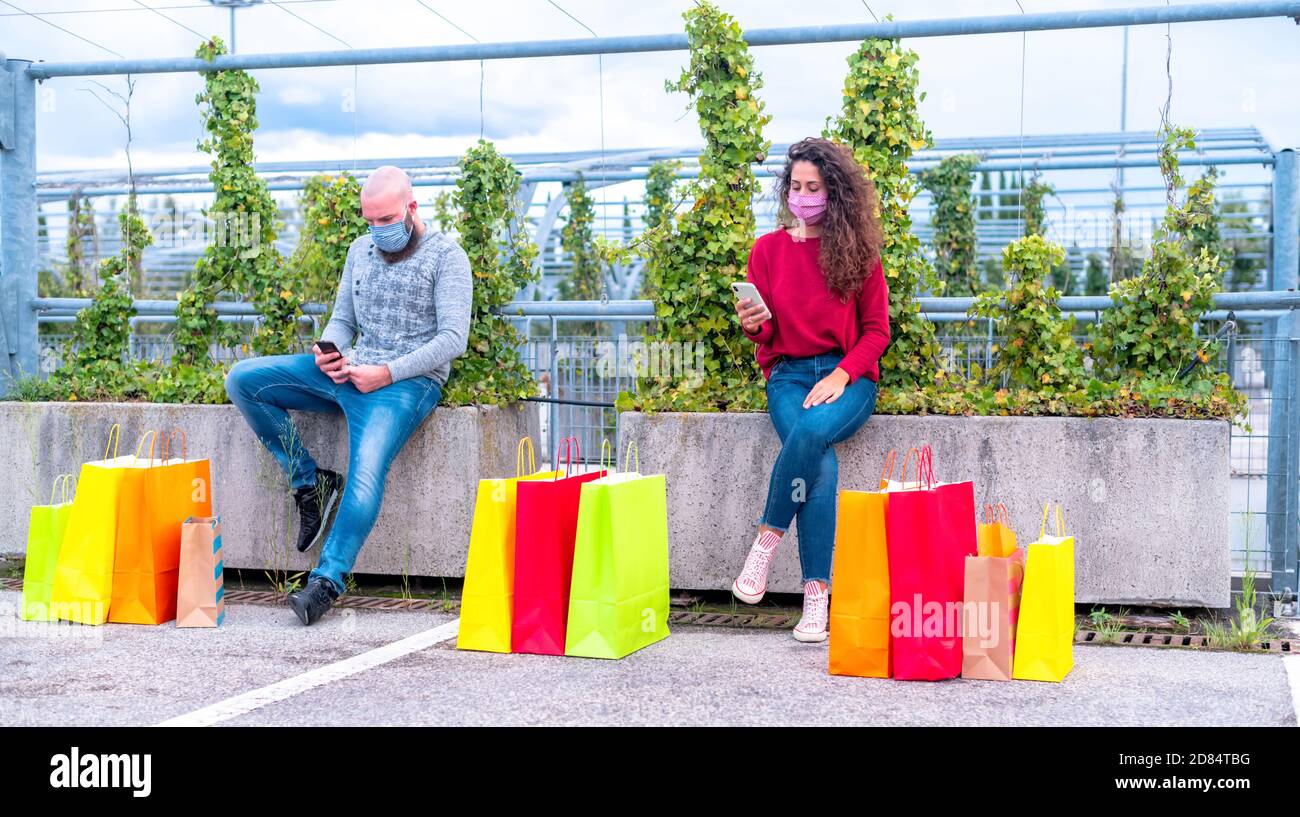 Après le shopping, heureux couple portant des masques de protection, assis sur un semoir en ville avec un smartphone en temps de coronavirus - les gens assis sur la ville être Banque D'Images