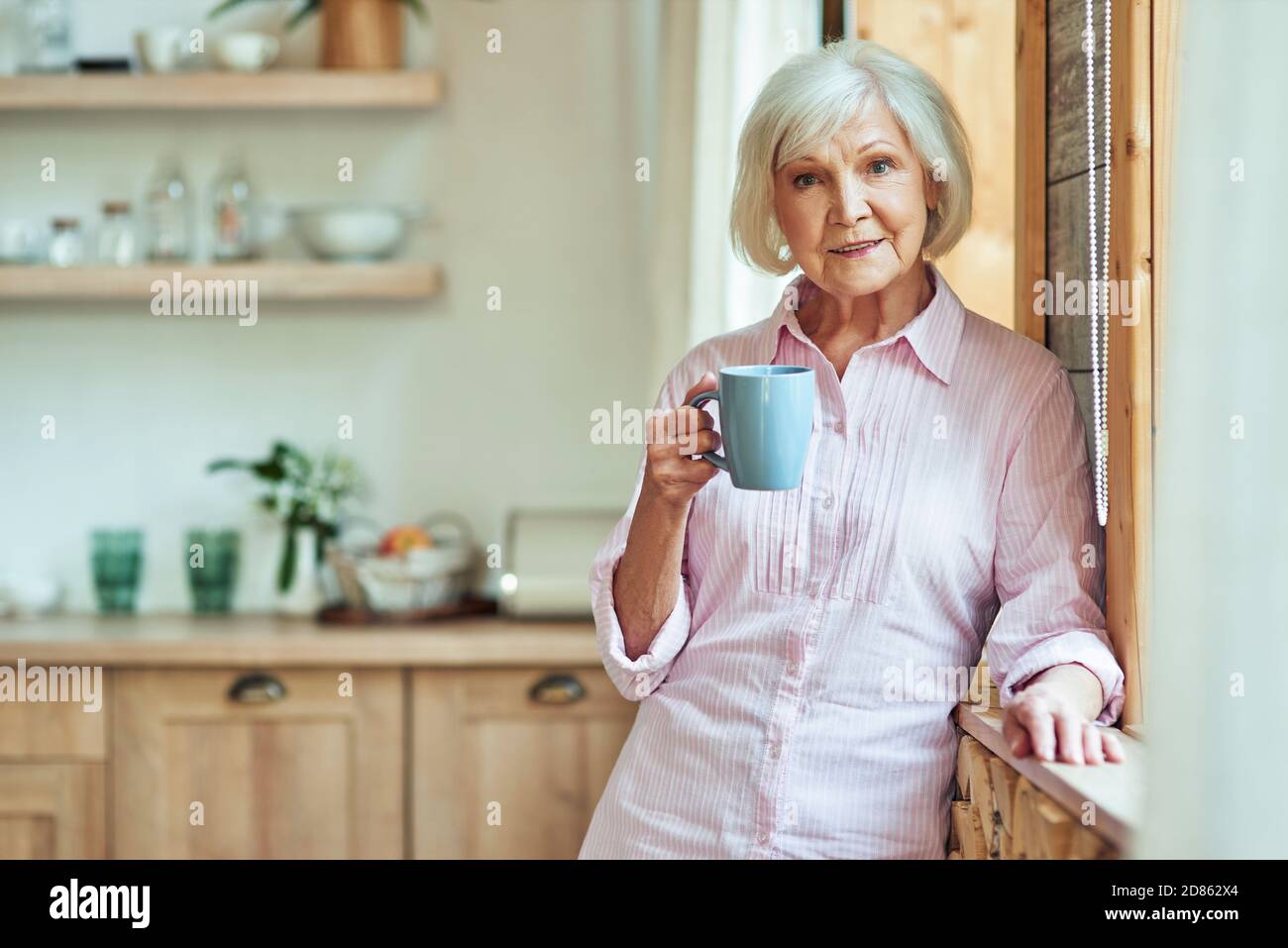 À la taille d'une femme élégante et souriante aux cheveux gris, debout près de la fenêtre et tenant une tasse de café dans la cuisine. Concept de style de vie domestique Banque D'Images